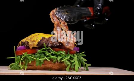 Handwerk Burger ist Kochen auf schwarzem Hintergrund. Bestehen: sauce, Rucola, Tomaten, Zwiebeln, Speck, Johannisbeeren Sauce, ricotta Käse, Brötchen und Fleisch. Stockfoto