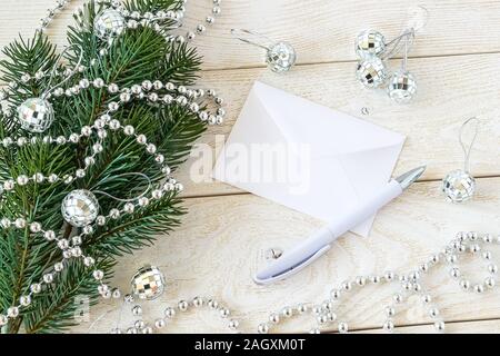 Weißen Umschlag und Kugelschreiber auf einem hölzernen Tisch in der Nähe eine Tanne Zweig mit silber Perle Girlande und Weihnachtskugeln Spiegel. Greeteing Karte für das Neue Jahr. Stockfoto