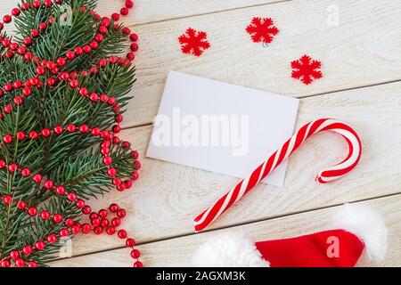 Leere greeteing Karte und sweet Candy Cane auf einen hölzernen Tisch in der Nähe eine Tanne Zweig mit roten Raupe Garland und Santa hat. Weihnachten und Neujahr Ferien. Stockfoto