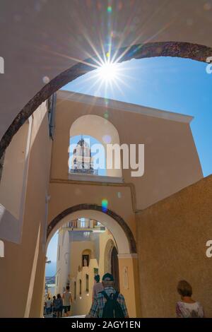 Santorini Griechenland - 8. August 2019; Sun blasts durch unter einem Bogen im Line up der architektonischen Merkmale der Bögen, Glockenturm und Uhr in Fira, der als Stockfoto