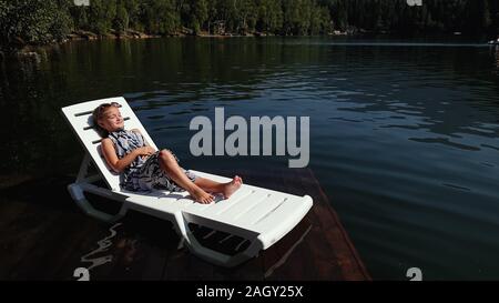 Kind Mädchen liegen auf der Sonnenliege in Sonnenbrille und Boho silk Schal. Kind auf einer Flut Holz unterwasser Pier. Das Pflaster wird mit Wasser im See bedeckt. Stockfoto