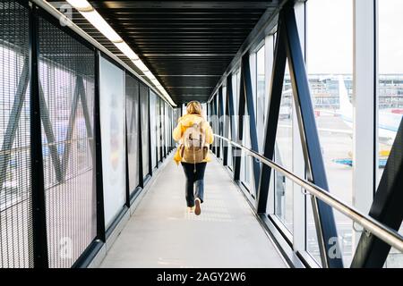 Ein kaukasischer blonde Frau geht durch den Flughafen mit gelber Jacke und Rucksack Stockfoto