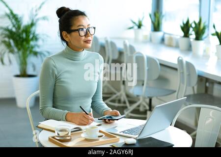 Hübsche asiatische Studenten in Brillen und lässige Pullover sitzen durch Tisch vor Laptop und Notizen in Notebook Stockfoto