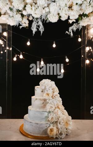 Weisse Hochzeitstorte mit Pfingstrosen und Rosen Blumen in der Nacht Zeremonie im Freien. Extra große dreistufige Hochzeitstorte mit weißen buttercream Stockfoto