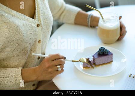 Die Hände der jungen Frau mit Cappuccino und Essen lecker Blueberry cheesecake beim Sitzen durch Tisch im Cafe Stockfoto