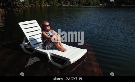 Kind Mädchen liegen auf der Sonnenliege in Sonnenbrille und Boho silk Schal. Kind auf einer Flut Holz unterwasser Pier. Das Pflaster wird mit Wasser im See bedeckt. Stockfoto