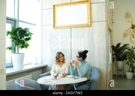 Zwei freundliche junge Frauen in der casualwear sitzen auf Weiche, bequeme Sofa von Tabelle in Café, Kaffee und Interagieren Stockfoto
