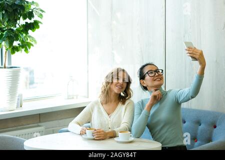 Zwei attraktive interkulturelle Mädchen in der casualwear Kaffee im Cafe und selfie während Freizeitaktivitäten genießen. Stockfoto