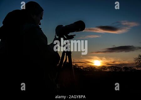 Der Fotograf Silhouette mit dem Tele Kamera holding Handy mit sichtbaren Datum (22. Dezember, Sonntag) gegen Winter Solstice Sonnenaufgang. Stockfoto