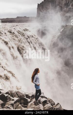 Junge Frau in Jeans und Shorts grau hoodie mit dramatischen Blick auf berühmte Island Wasserfall Dettifoss. Atemberaubende Landschaft, Strom von Wasser, mächtigsten Wasserfall Europas. Stockfoto