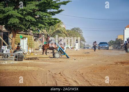Mbour SENEGAL - Apr 26, 2019: Unbekannter senegalesische Frau mit Baby sind zu Fuß eine staubige Straße mitten in der Stadt. Es gibt ein Auto auf dem Stockfoto