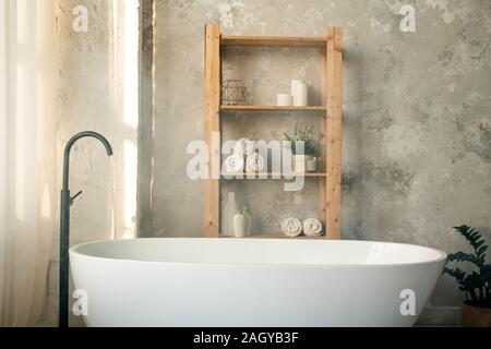 Große Porzellan weiße Wanne und Holzregale gegen graue Wand im Bad Stockfoto