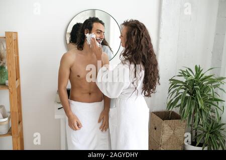 Junge Frau im weißen Bademantel Anwendung Rasierschaum auf Bart von ihrem Mann Stockfoto
