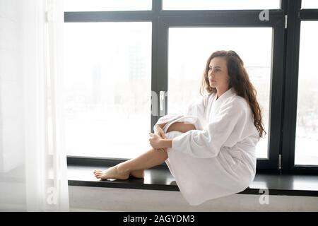 Junge brunette heitere Frau in weichen, weißen Bademantel entspannen auf Fensterbank Stockfoto