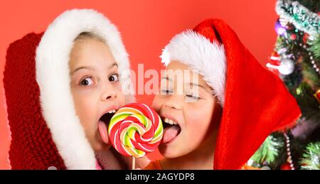 Mädchen beißen Lutscher, zusammen zu schließen. Schwestern in Weihnachten Hüte mit Geschenkboxen offen präsentiert. Feiern und Urlaub Konzept. Kinder mit froh Gesichter essen Süßigkeiten auf rotem Hintergrund. Stockfoto