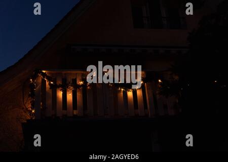 Weihnachten Kränze mit Kerzenlicht auf hölzernen Balkon vor einem Haus in der Dämmerung in der Weihnachtszeit. Baum und Haus als eine Form. Low Angle View. Stockfoto