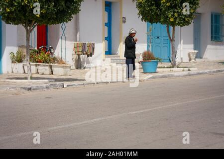 Ein Mann mit Hut steht mitten am Tag vor dem Haus in der tunesischen Stadt Hergla. Die Straße ist von Sonnenlicht durchflutet. Stockfoto