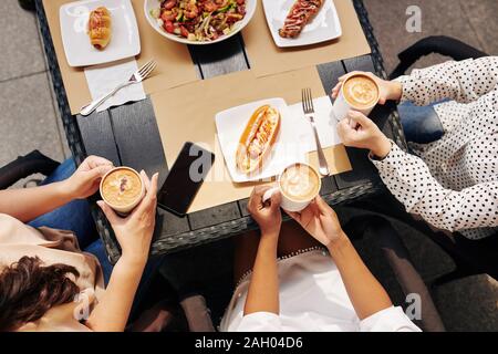 Zugeschnittenes Bild der weiblichen Freunde essen lecker Essen und Trinken große Tassen Cappuccino im Café draußen, Ansicht von oben Stockfoto
