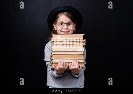 Fröhliche Schülerin mit toothy Lächeln holding Stapel von Büchern beim Stehen in der Isolation auf schwarzem Hintergrund Stockfoto