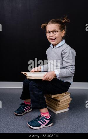 Adorable Schülerin in Brillen und Casualwear sitzen auf Stapel Bücher aufgeregt, während einer von ihnen lesen vor der Kamera gegen die Tafel Stockfoto