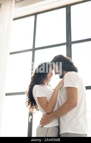 Junge zärtlich lächelnd Paar in den weißen T-Shirts gegen grosse Fenster schließen einander Stockfoto