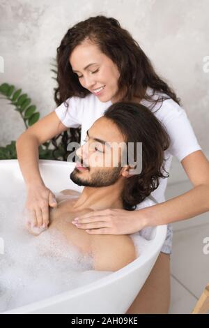 Hübsche junge lächelnde Frau, die Massage der Brust zu ihrem Mann in Badewanne mit heißem Wasser und Schaum Stockfoto