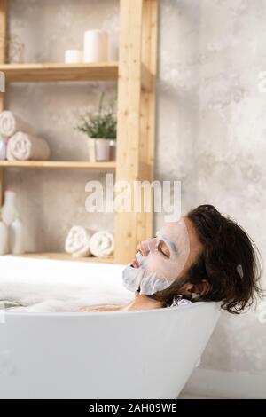 Entspannt junger Mann mit Maske, während in der Badewanne mit heißem Wasser und Schaum liegen Stockfoto