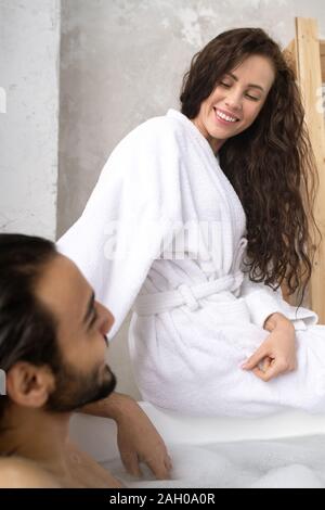 Glückliche junge lächelnde Frau in weißen Bademantel im Gespräch mit ihrem Mann in Badewanne Stockfoto