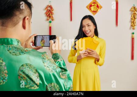 Junger Mann fotografieren schöne Freundin in traditioneller Kleidung posiert mit gelben Glück Geld Umschläge an TET-Fest Stockfoto