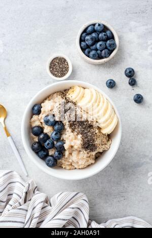 Haferflocken Brei in der Schüssel mit Blaubeeren, Banane und Chia Samen auf grauem Beton Hintergrund, gesundes Frühstück essen. Ansicht von oben. Vertikale Zusammensetzung Stockfoto