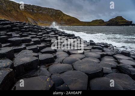 Meer Wellen schlagen die natürliche sechseckige Steine an der Küste namens Giant's Causeway, ein Wahrzeichen in Nordirland, die Klippen, die in der Umgebung des Stockfoto