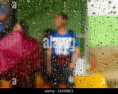 Regentropfen auf Fenster - Pendler mit Sonnenschirmen an der Bushaltestelle warten, von einem Auto, das Fenster auf der Beifahrerseite. Zur Veranschaulichung ein regnerischer Tag/nasses Wetter/gefangen Ich Stockfoto