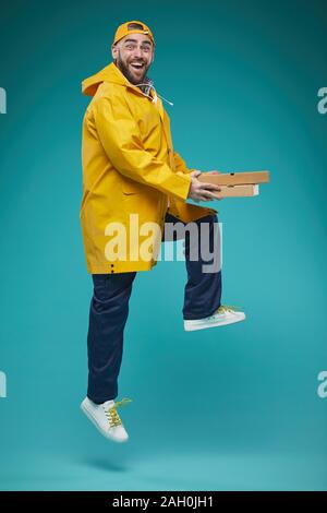 Studio voller Schuß von fröhlichen Food Delivery Man tragen gelbe Uniform springen mit pizzaa Boxen in Händen Stockfoto