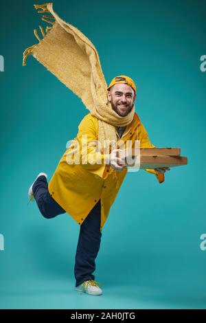 Vertikale Studio voller Schuß von jungen kaukasischen Mann tragen gelbe Uniform liefern Pizza, blau türkis Hintergrund Stockfoto