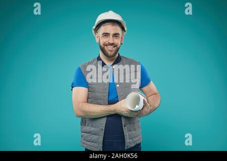 Horizontale medium Portrait von gutaussehenden jungen Mann in Uniform und schützende Kopfbedeckung holding Bauplan in die Kamera lächeln Stockfoto