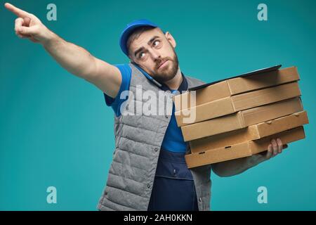 Horizontale medium Studio shot von Lebensmitteln Lieferung männlichen Arbeitnehmers holding Pizzakartons, am Telefon sprechen und zeigte auf etwas Stockfoto