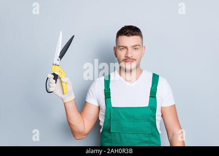 Foto von Stattlichen starken Kerl Haltearm Gartenarbeit Schere hören Kundenauftrag über schneiden Sträucher tragen grüne Latzhose grau isoliert Hintergrund Stockfoto