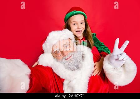 Nahaufnahme Foto von redhair elf in grünen Hut Kopfbedeckung und Santa Claus mit weißem Haar, selfie v-Zeichen tragen Brillen Brillen über Rot isoliert Stockfoto