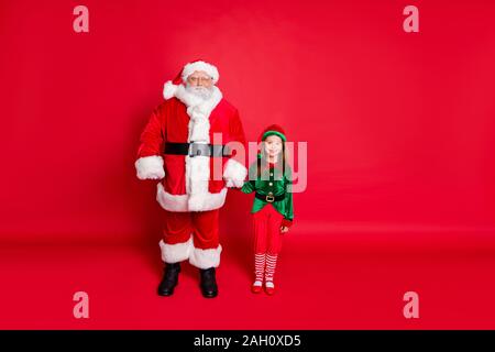 Volle Größe Foto von fröhlichen zwei Santa und elf halten sich an den Händen bereit Neujahr noel tragen helle grüne Kostüm über den roten Hintergrund isoliert zu genießen Stockfoto