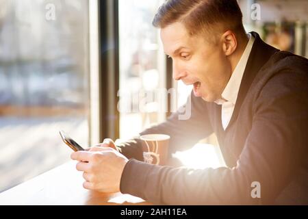 Überrascht Mann mit Handy in der Hand sitzen in der Nähe der Fenster im Cafe bei Tag Stockfoto