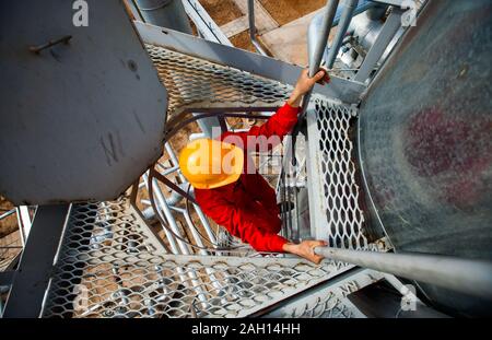 Arbeitnehmer in rot Arbeit tragen und gelben Helm auf grauem Raster auf Ölraffinerie Anlage. Stockfoto