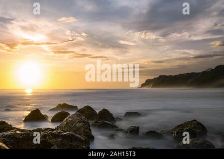 Einen atemberaubenden Blick auf die felsige Küste durch gleichmäßige, seidige Meer bei Sonnenuntergang getaucht. Melasti Beach mit seinen Klippen in der Ferne, in Bali, Indonesien. Stockfoto