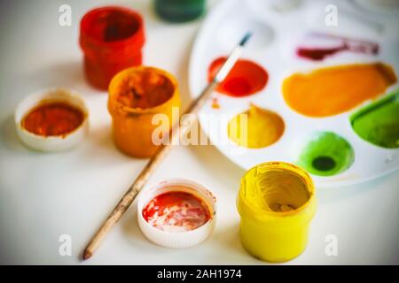 Auf einem weißen Tisch Es gibt Kunst liefert - ein weißes Plastik Palette zum Mischen von Farben, verschiedene Farben von gouache Farbe, Pinsel und einen Deckel von einem Glas Stockfoto