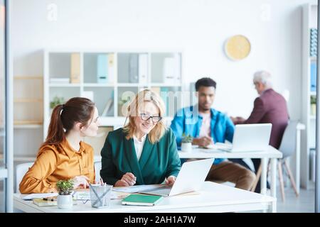 Zwei Geschäftsfrauen sitzen am Tisch und diskutieren online Präsentation im Team mit anderen Menschen im Hintergrund arbeiten Stockfoto