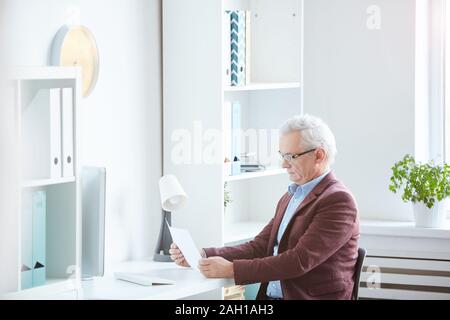Schwere reifen Geschäftsmann in Brillen an seinem Arbeitsplatz sitzen und lesen ein business Vertrag im modernen Büro Stockfoto