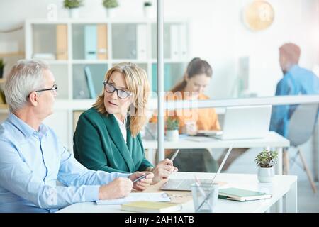 Reife Geschäftsfrau in Brillen im Gespräch mit den Reifen Geschäftsmann, während Sie am Schreibtisch sitzt mit Laptop im Büro Stockfoto