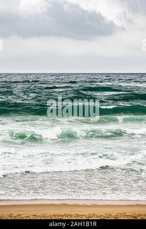 Raue Meer und Wellen in zum Strand und Felsen im Vordergrund, Surfer im Hintergrund Stockfoto