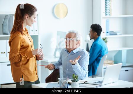 Männliche reife Führer sitzt an seinem Arbeitsplatz und im Gespräch mit den jungen Geschäftsfrau im Büro Stockfoto