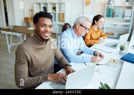 Junge afrikanische Unternehmer Lächeln auf die Kamera, während auf Laptop am Tisch zusammen mit seinen Kollegen Stockfoto