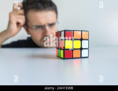 Mann mit Brille verwirrten Blick auf der Rubik's Cube seinen Kopf kratzen, ohne zu wissen, wie es zu lösen Stockfoto